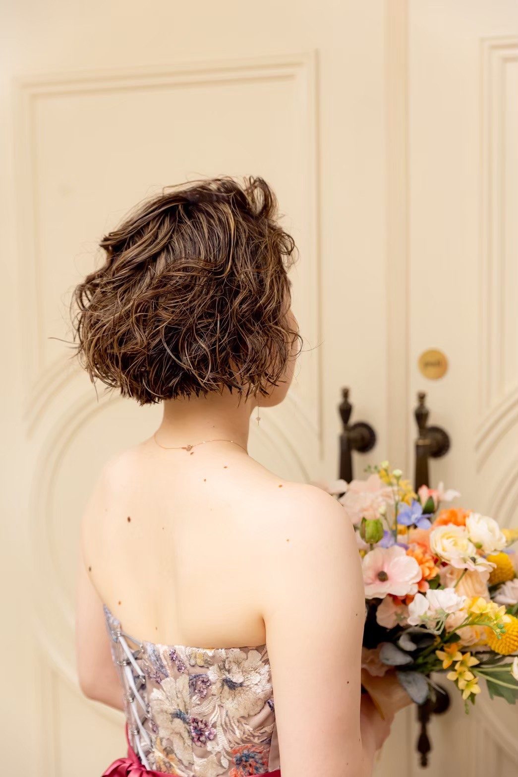 カット後のボブスタイルで、ブーケを手に会場の扉の前に立つ花嫁の後ろ姿